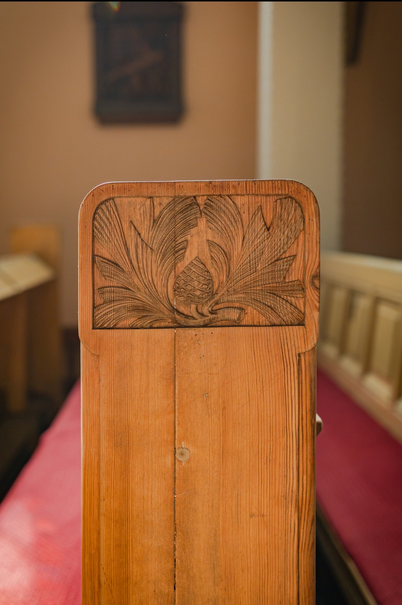 Drewniana ławka kościelna z rzeźbionym motywem roślinnym, widoczny fragment czerwonego obicia siedziska, w tle zarys innego mebla.