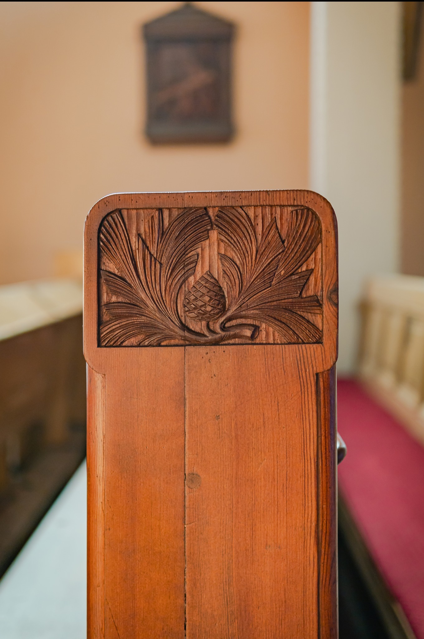 Drewniana ławka kościelna z rzeźbionym motywem szyszki i liści akantu w górnej części oparcia, widoczne detale snycerskie i ciepła barwa drewna.