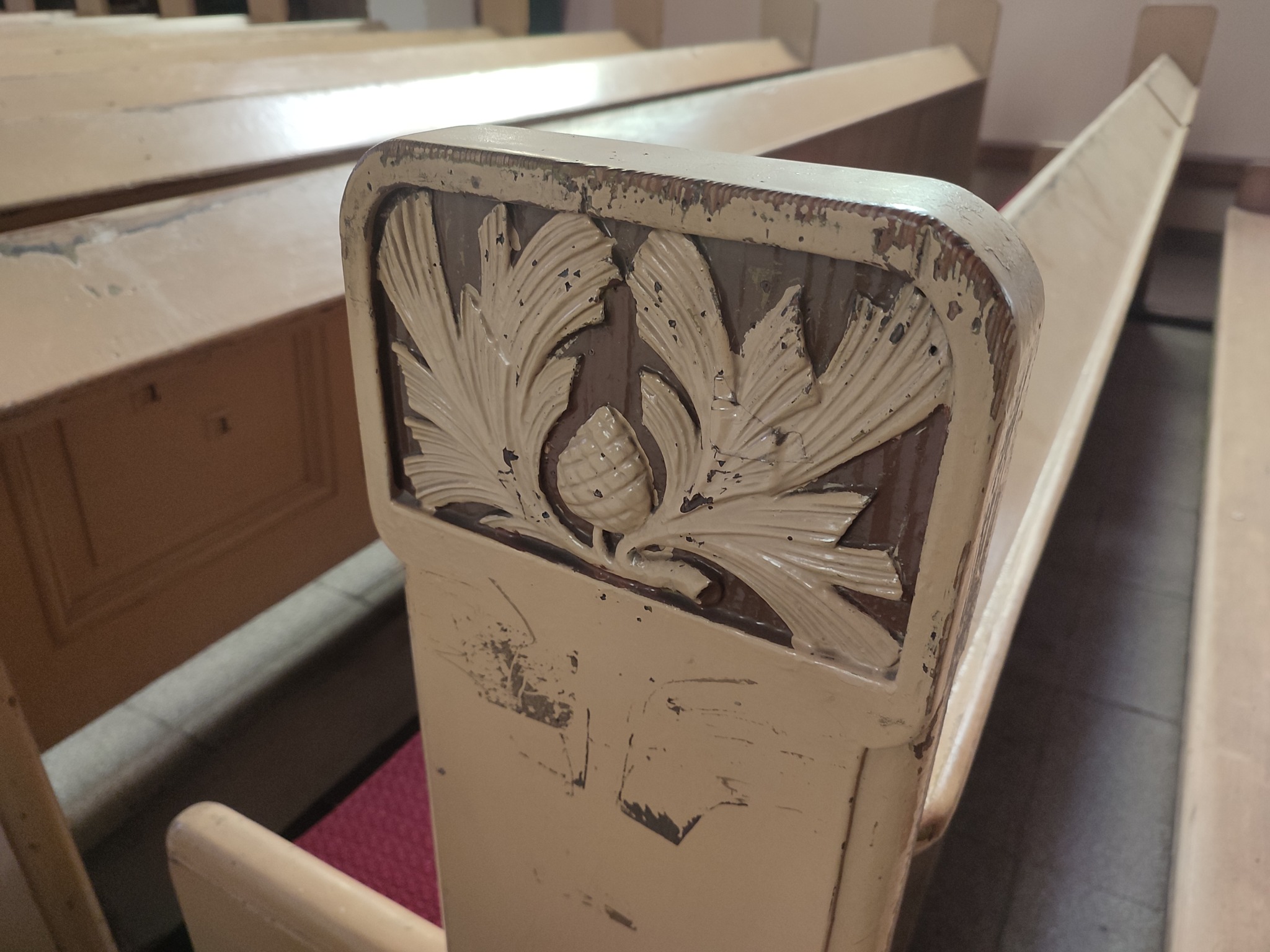 Detal zdobionego, odrapanego oparcia ławki kościelnej z motywem roślinnym i szyszki na tle rzędów ławek.