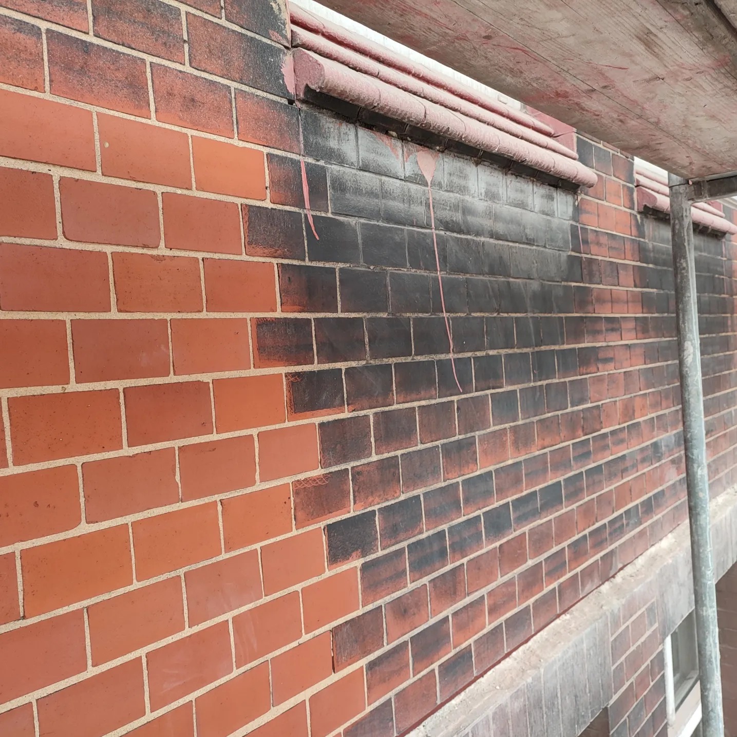 Częściowo wyczyszczona ceglana elewacja budynku z widocznymi różnicami w kolorze cegieł, z fragmentem rusztowania po prawej stronie.