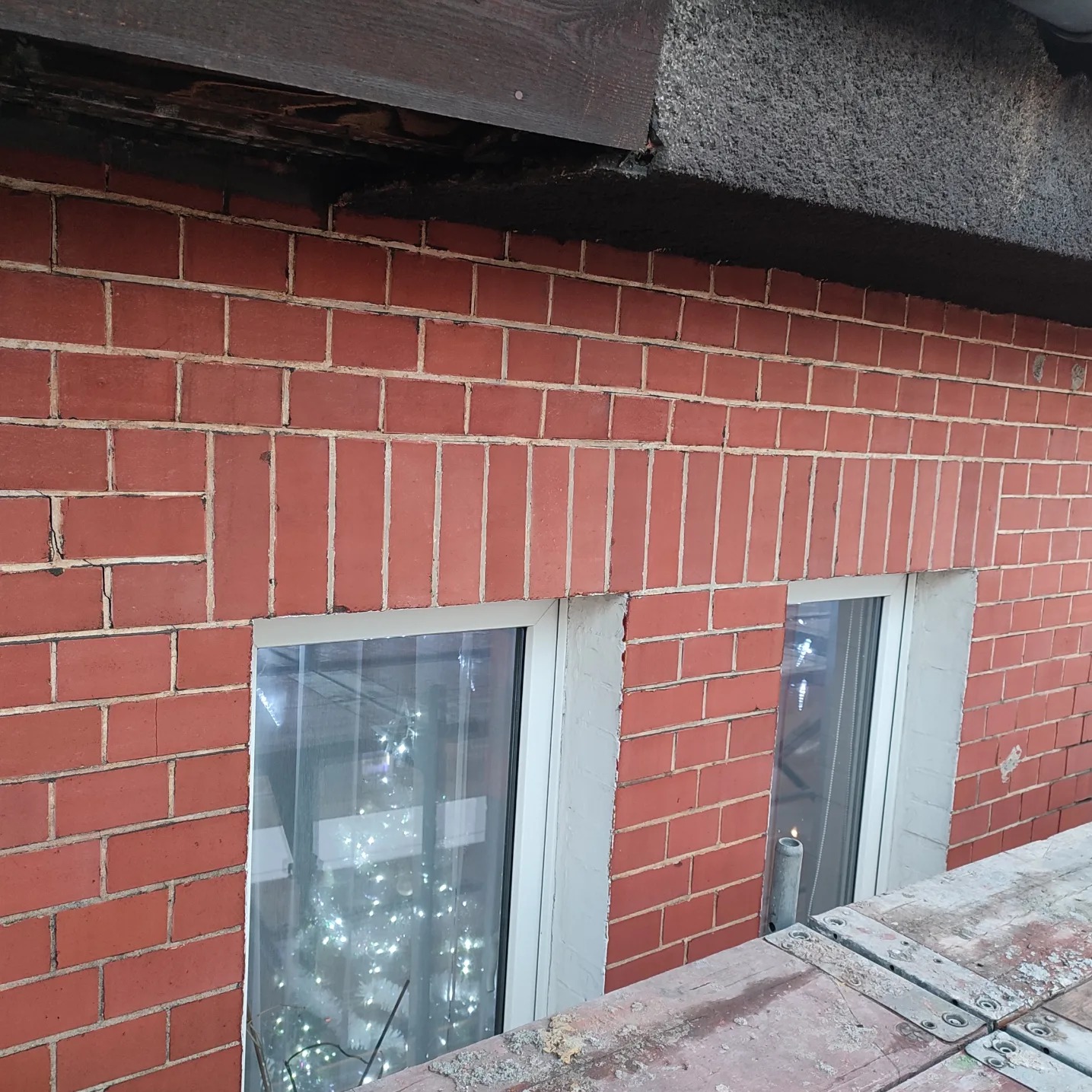 Czerwona ceglana ściana budynku z dwoma oknami z białymi ramami, widok z rusztowania, w oknach widoczne dekoracje świetlne.