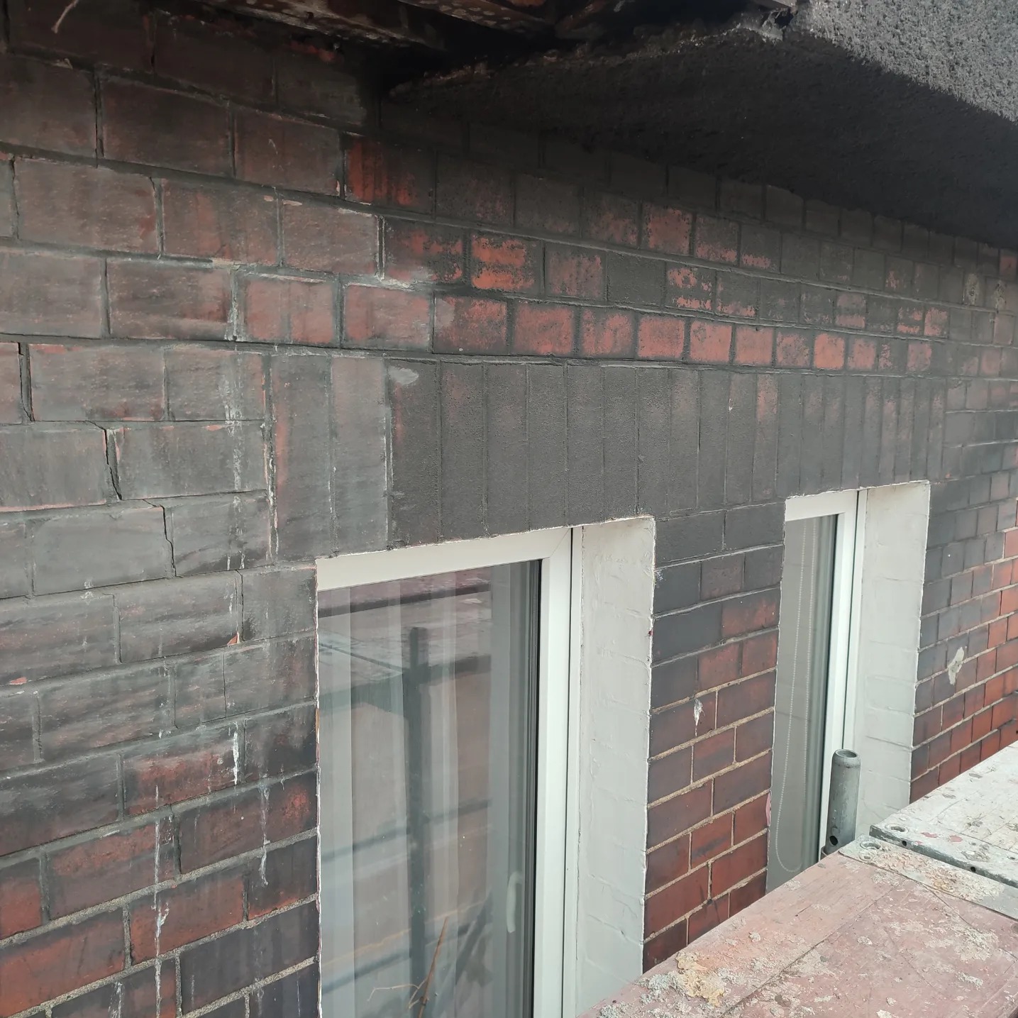 Częściowo wyczyszczona ceglana elewacja budynku z dwoma oknami z białymi ramami, widoczne ślady starego brudu i porostów, rusztowanie w dolnym rogu.