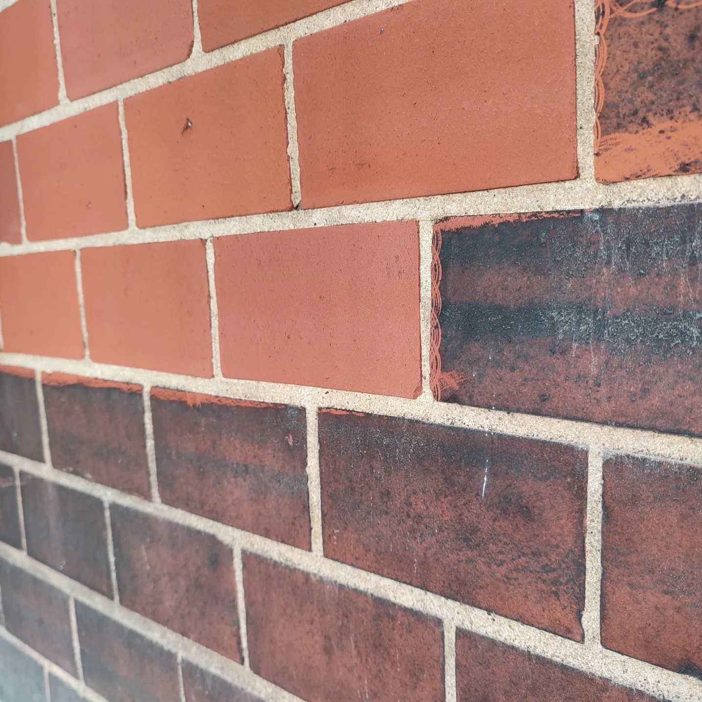Fragment muru z cegły klinkierowej w dwóch odcieniach czerwieni, z widocznymi śladami zabrudzeń na niektórych cegłach i jasną fugą, sfotografowany pod kątem.