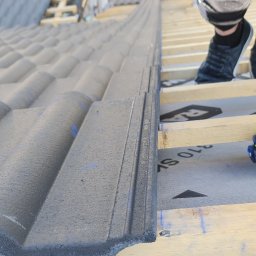 Pro Dach - Układanie szarej dachówki na drewnianej konstrukcji dachu, widoczne buty dekarza i membrana dachowa.