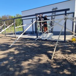 Pro Dach - Montaż konstrukcji z czarnych belek przy białym budynku, z użyciem drewnianych podpór i drabiny, w słoneczny dzień.