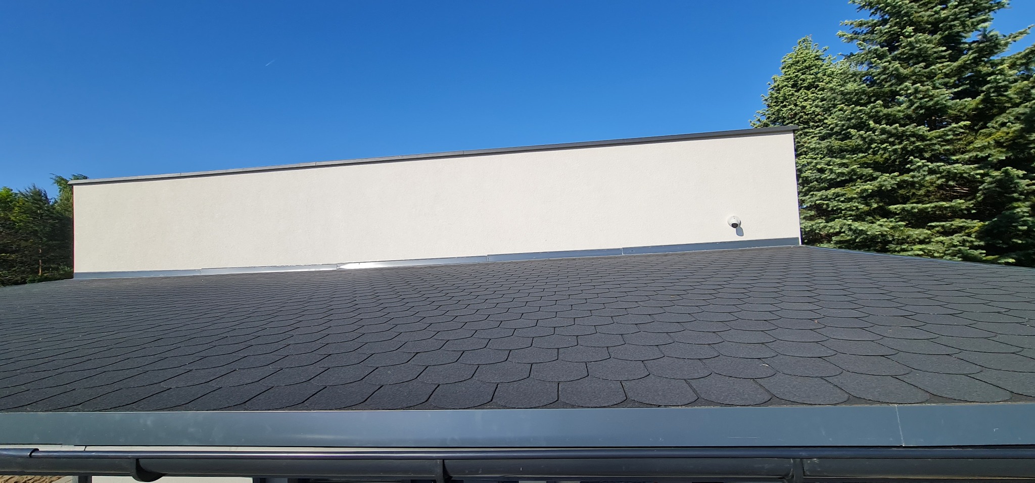 Dach pokryty czarnym gontem bitumicznym w kształcie łuski, z widoczną obróbką blacharską i fragmentem elewacji budynku oraz rynną w kolorze szarym, na tle błękitnego nieba i zielonych drzew.