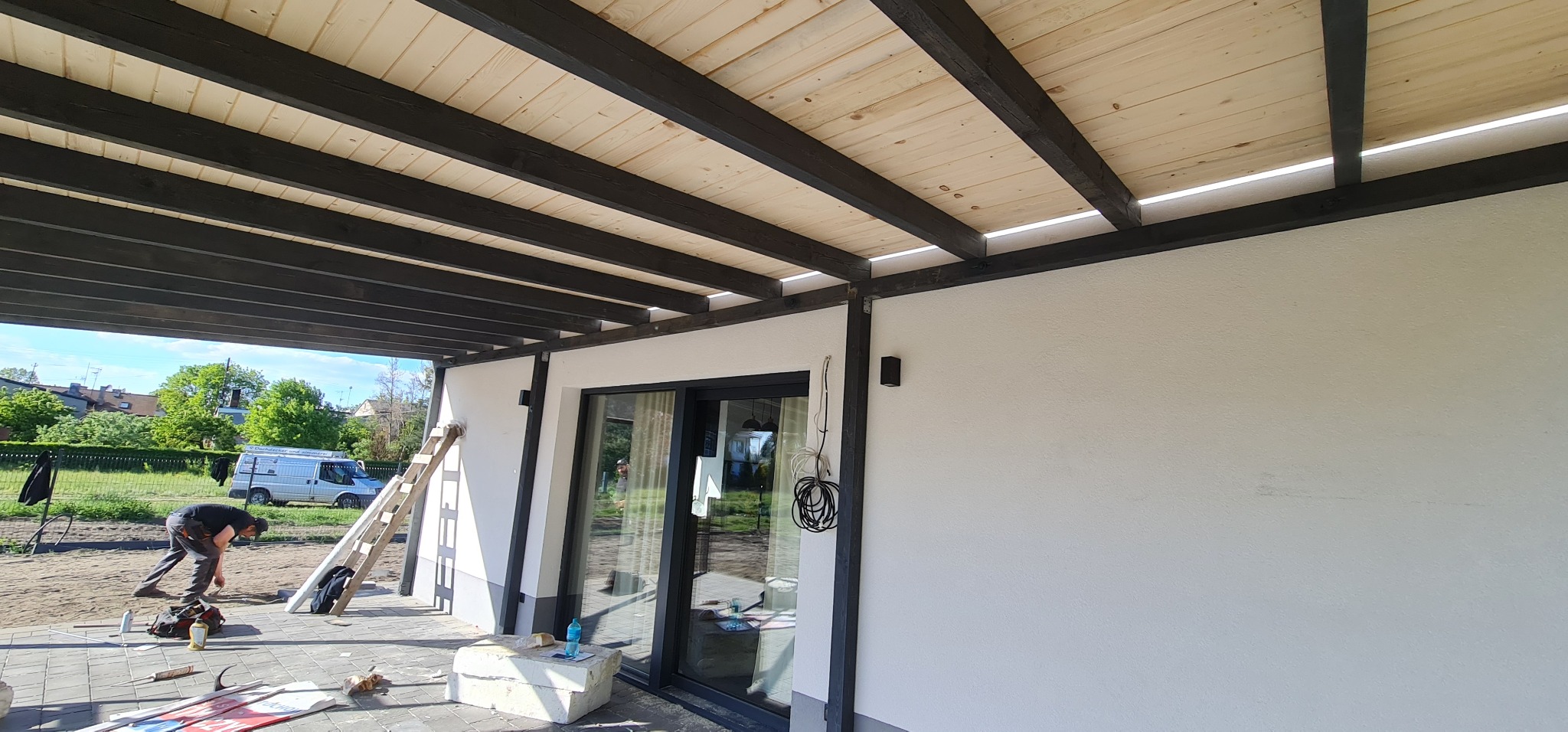Drewniana pergola z czarnymi belkami i jasnym deskowaniem, widok z zewnątrz, w tle pracownik układający kostkę brukową.