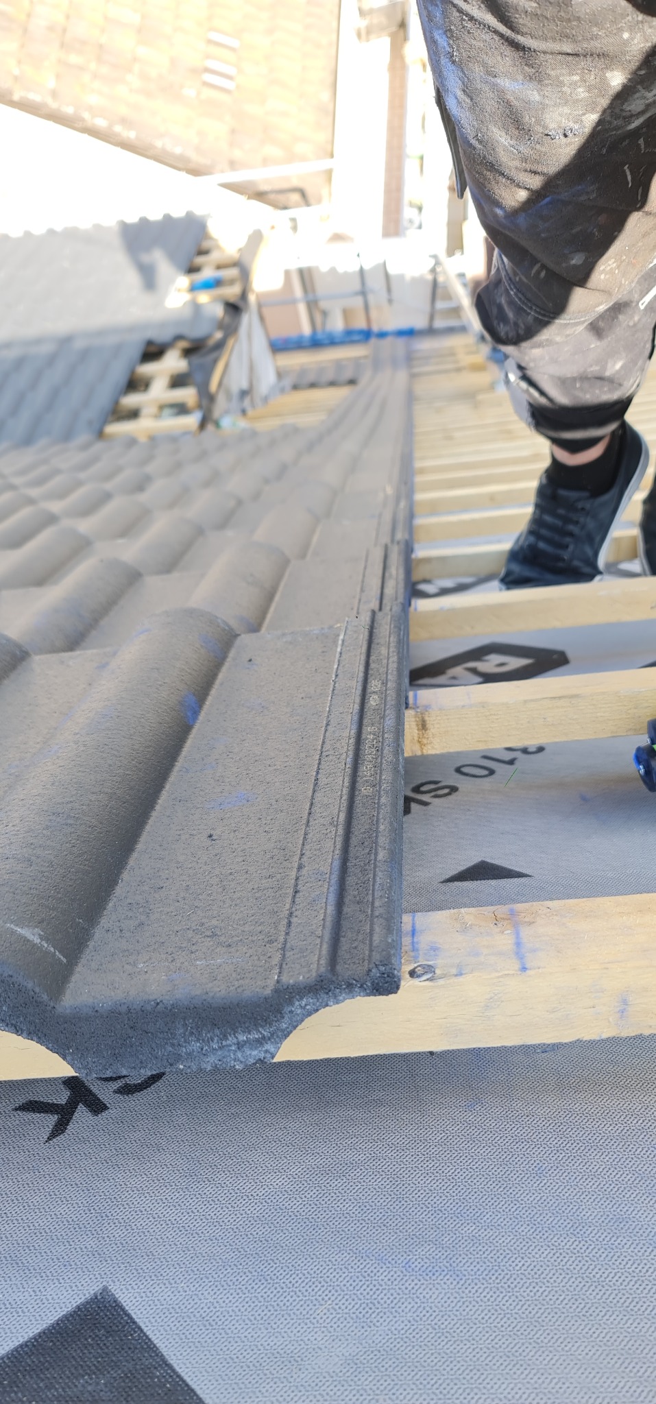 Układanie szarej dachówki na drewnianej konstrukcji dachu, widoczne buty dekarza i membrana dachowa.