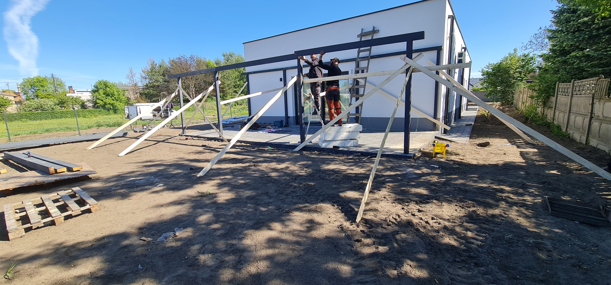 Montaż konstrukcji z czarnych belek przy białym budynku, z użyciem drewnianych podpór i drabiny, w słoneczny dzień.