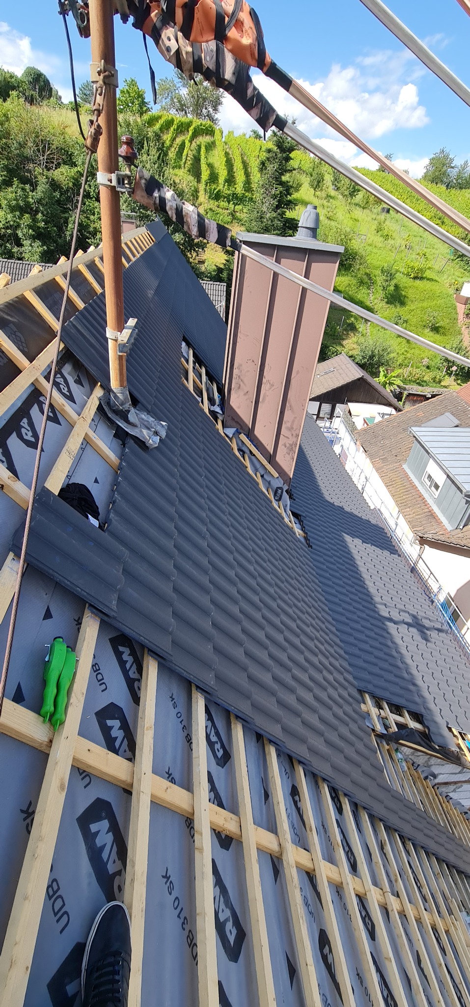 Montaż dachówki ceramicznej w kolorze grafitowym na drewnianej konstrukcji dachu, widoczna membrana dachowa, komin w trakcie obróbki blacharskiej i elementy rusztowania na tle zielonych wzgórz.