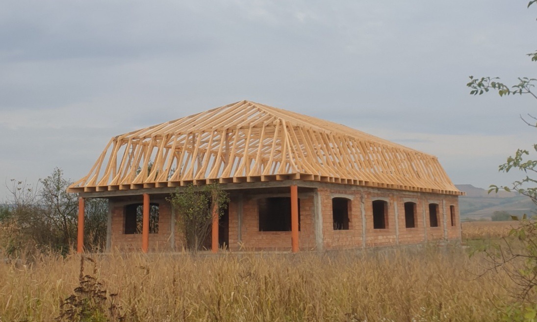 Surowa konstrukcja domu z cegły z drewnianą więźbą dachową w trakcie budowy, widok z zewnątrz na tle polnego krajobrazu.