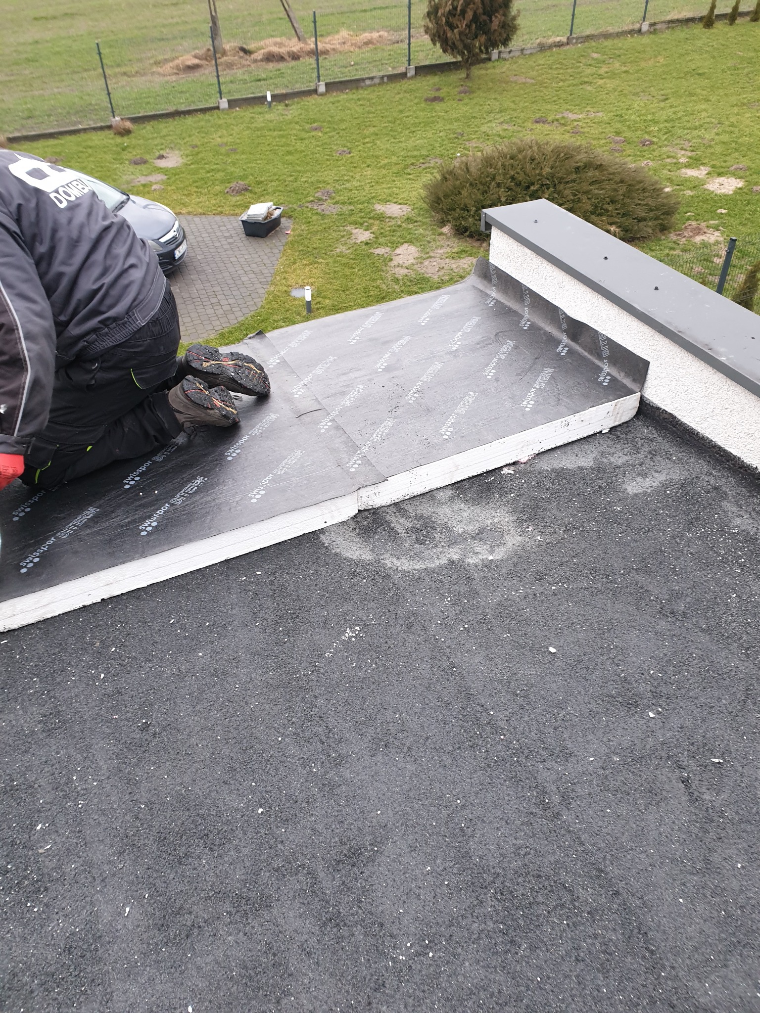 Dekarz na kolanach układa warstwę izolacyjną z czarnej membrany Swissport Biterm na dachu budynku z płaskim dachem i attyką.