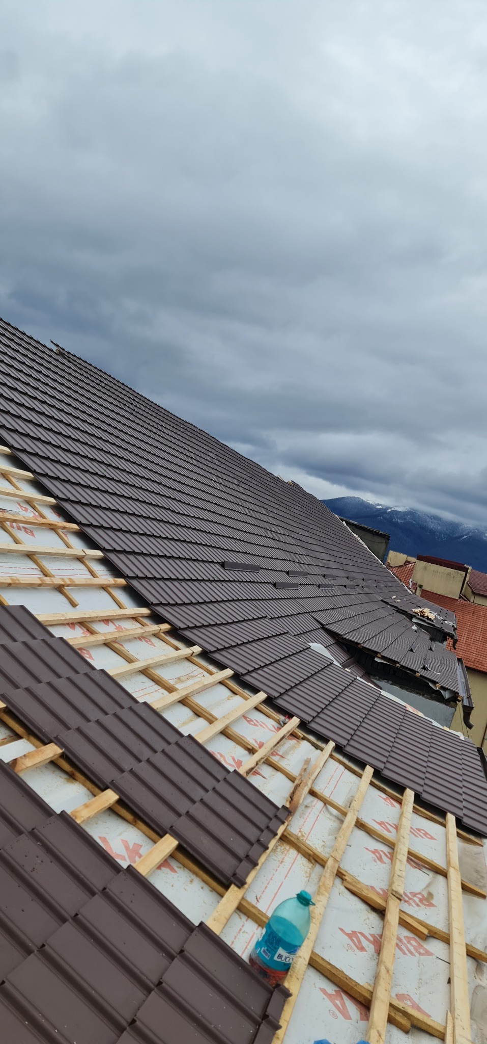 Montaż brązowej dachówki na drewnianej konstrukcji, widoczne ocieplenie, w tle pochmurne niebo i góry.