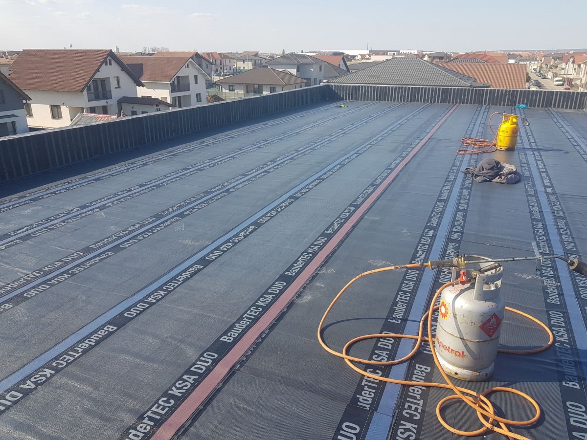 Hydroizolacja dachu płaskiego z papy termozgrzewalnej BauderTEC KSA DUO, widoczne butle gazowe i palniki dekarskie, w tle zabudowa jednorodzinna.