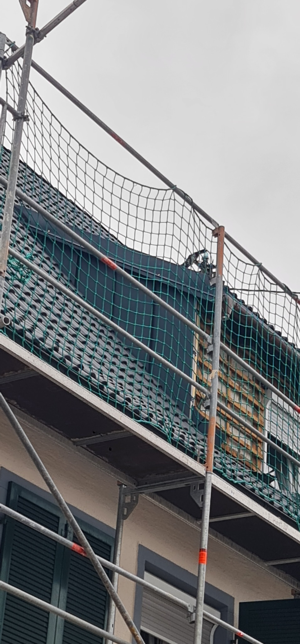 Fragment dachu z czarną dachówką widziany z poziomu rusztowania zabezpieczonego siatką, okno z drewnianą ramą w tle.