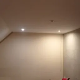 Poddasze po remoncie, ściany wygładzone i pomalowane na jasny beż, widoczne dwa punkty oświetleniowe w suficie, fragment ściany z cegły po lewej stronie.