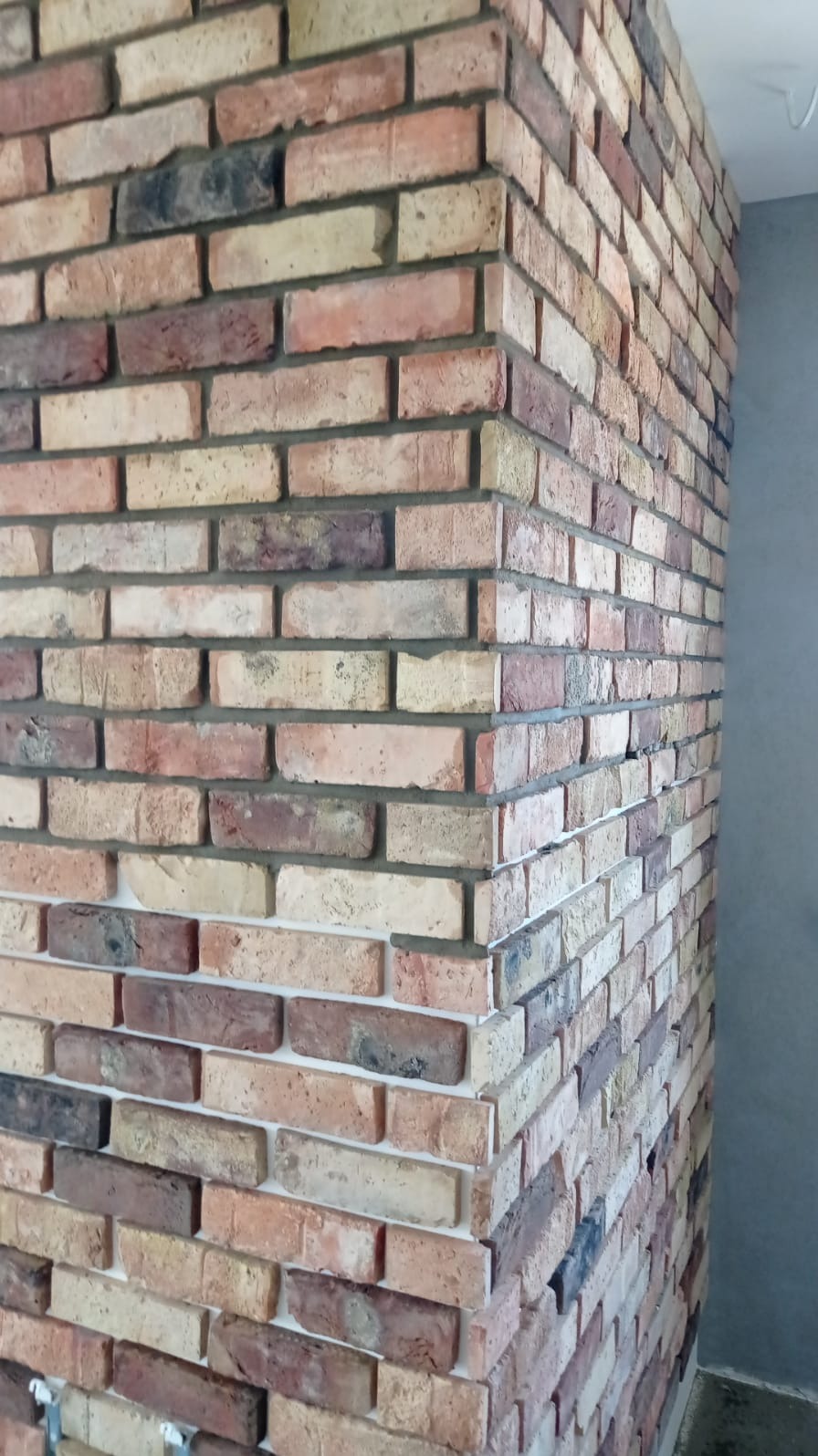 Dekoracyjna ściana z cegieł klinkierowych w odcieniach beżu, brązu i czerwieni, tworząca narożnik w pomieszczeniu z szarą ścianą w tle.