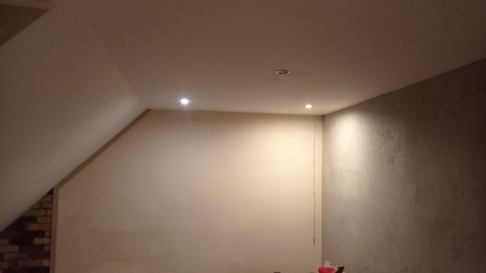 Poddasze po remoncie, ściany wygładzone i pomalowane na jasny beż, widoczne dwa punkty oświetleniowe w suficie, fragment ściany z cegły po lewej stronie.