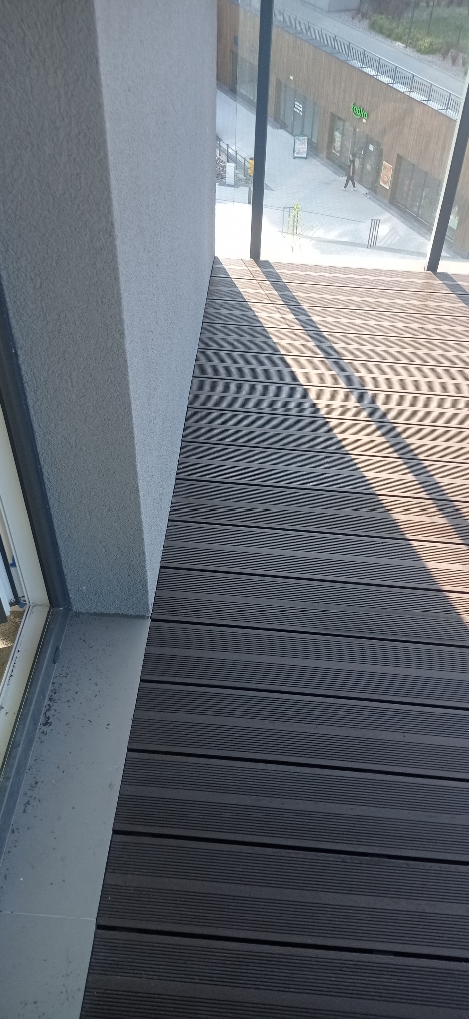 Szare deski tarasowe ułożone na balkonie, widok z góry, cień rzucany przez barierkę, widok na plac z budynkami w tle.