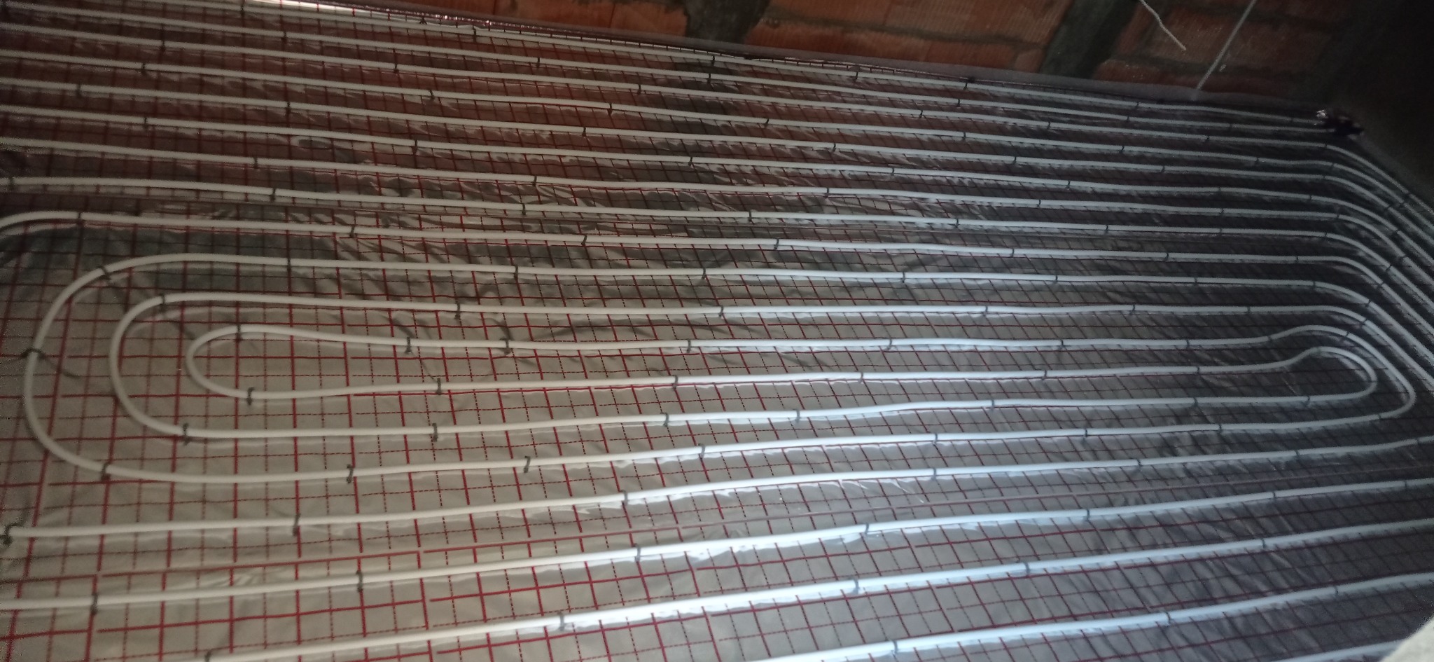 Rozłożone rury ogrzewania podłogowego na folii z czerwoną siatką, widoczne złącza mocujące rury do podłoża, surowy strop w tle.