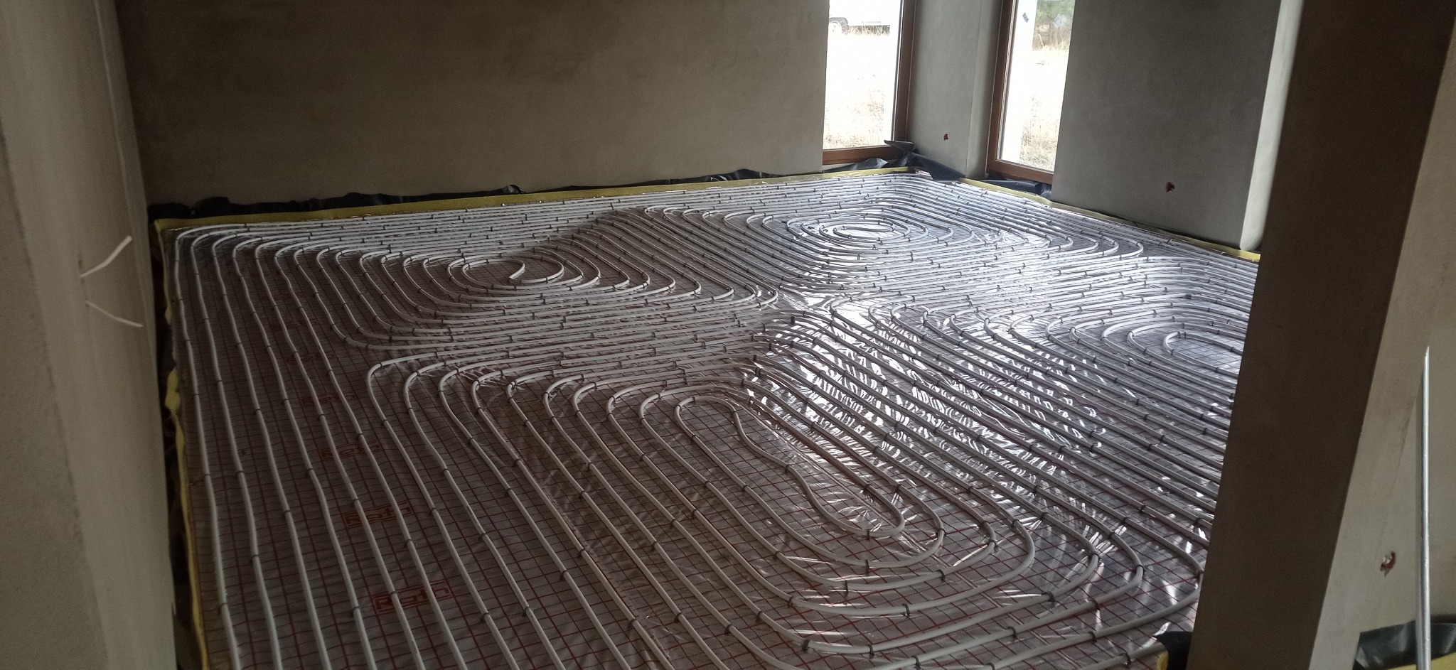 Instalacja ogrzewania podłogowego z rur na podkładzie z siatki w surowym wnętrzu z oknami.