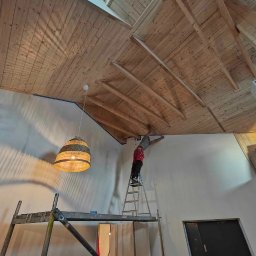 Mariusz Kowalski - Pracownik na drabinie maluje ścianę przy drewnianym suficie z oknem dachowym, widoczne rusztowanie i wisząca lampa.