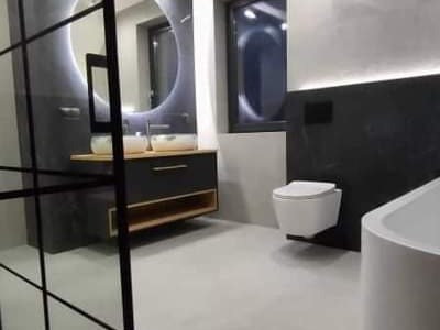 Nowoczesna łazienka z okrągłym lustrem podświetlanym LED, wiszącą toaletą i czarną ścianą akcentującą, widziana zza szklanej ścianki prysznicowej.