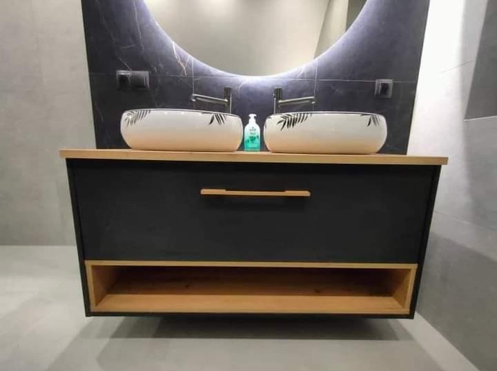 Nowoczesna łazienka z podwójną umywalką nablatową w geometryczne wzory, zamontowaną na czarnej szafce z drewnianym blatem i otwartą półką; nad umywalkami podświetlane lustro w kształcie łuku.