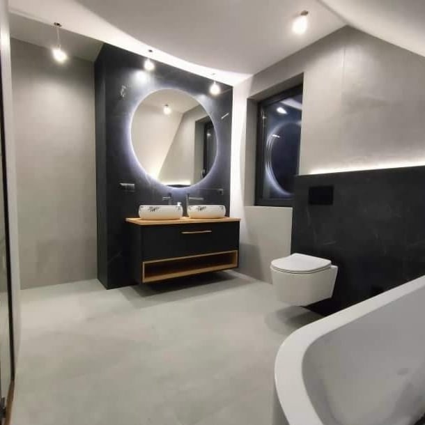Elegancka łazienka z okrągłym lustrem podświetlanym LED, dwiema umywalkami nablatowymi w geometryczne wzory, czarną szafką z drewnianymi akcentami i minimalistyczną toaletą podwieszaną.