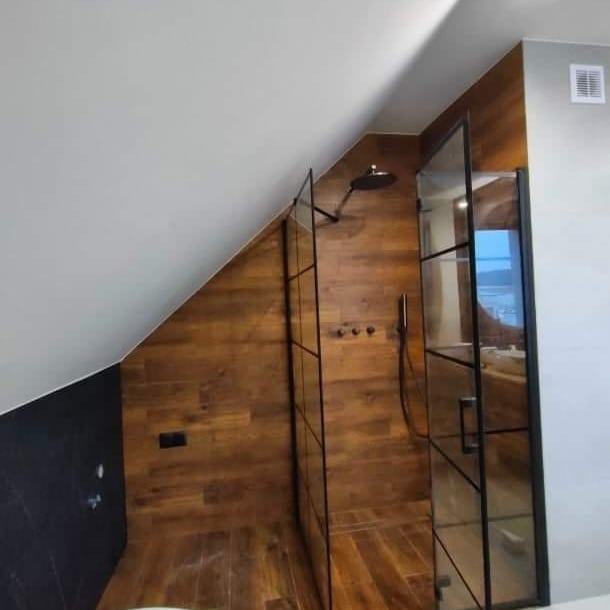 Nowoczesna kabina prysznicowa w łazience na poddaszu, z drewnopodobnymi panelami na ścianach i czarnymi, metalowymi ramami szklanych drzwi.