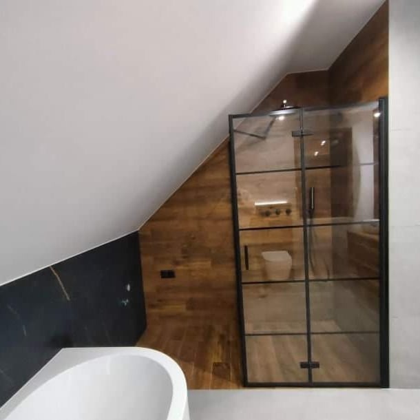 Nowoczesna łazienka na poddaszu ze skośnym sufitem, kabina prysznicowa z czarnymi szprosami, drewnopodobne płytki ścienne i wanna narożna.