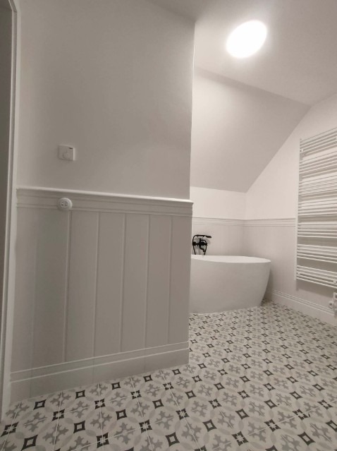 Nowoczesna łazienka w odcieniach bieli z wolnostojącą wanną i wzorzystą podłogą, widok z perspektywy wejścia, akcent na panel ścienny z dekoracyjną listwą.