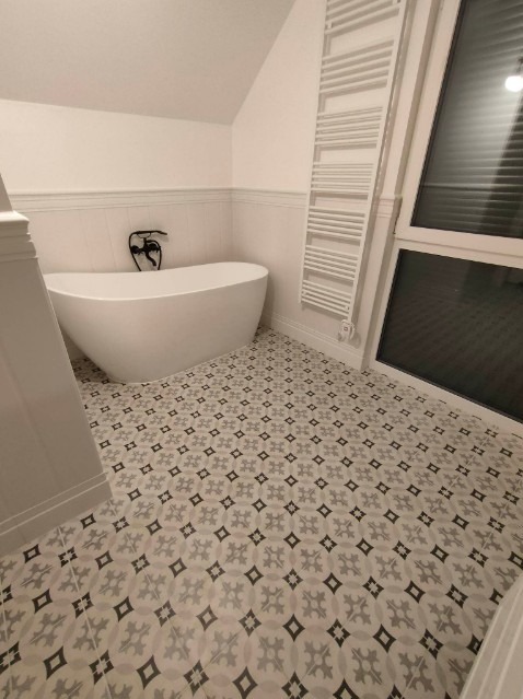 Nowoczesna łazienka z wolnostojącą białą wanną, czarną armaturą i grzejnikiem drabinkowym, wykończona płytkami podłogowymi w geometryczny wzór.