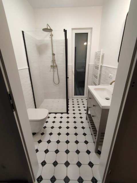 Nowoczesna łazienka z czarno-białą podłogą w karo, kabiną prysznicową z czarną ramą i złotą armaturą, oraz szarą szafką pod umywalką.