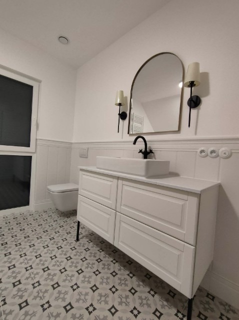 Nowoczesna łazienka z białą szafką pod umywalkę na czarnych nóżkach, prostokątną umywalką, czarną baterią, lustrem w złotej ramie i wzorzystą szaro-białą podłogą.