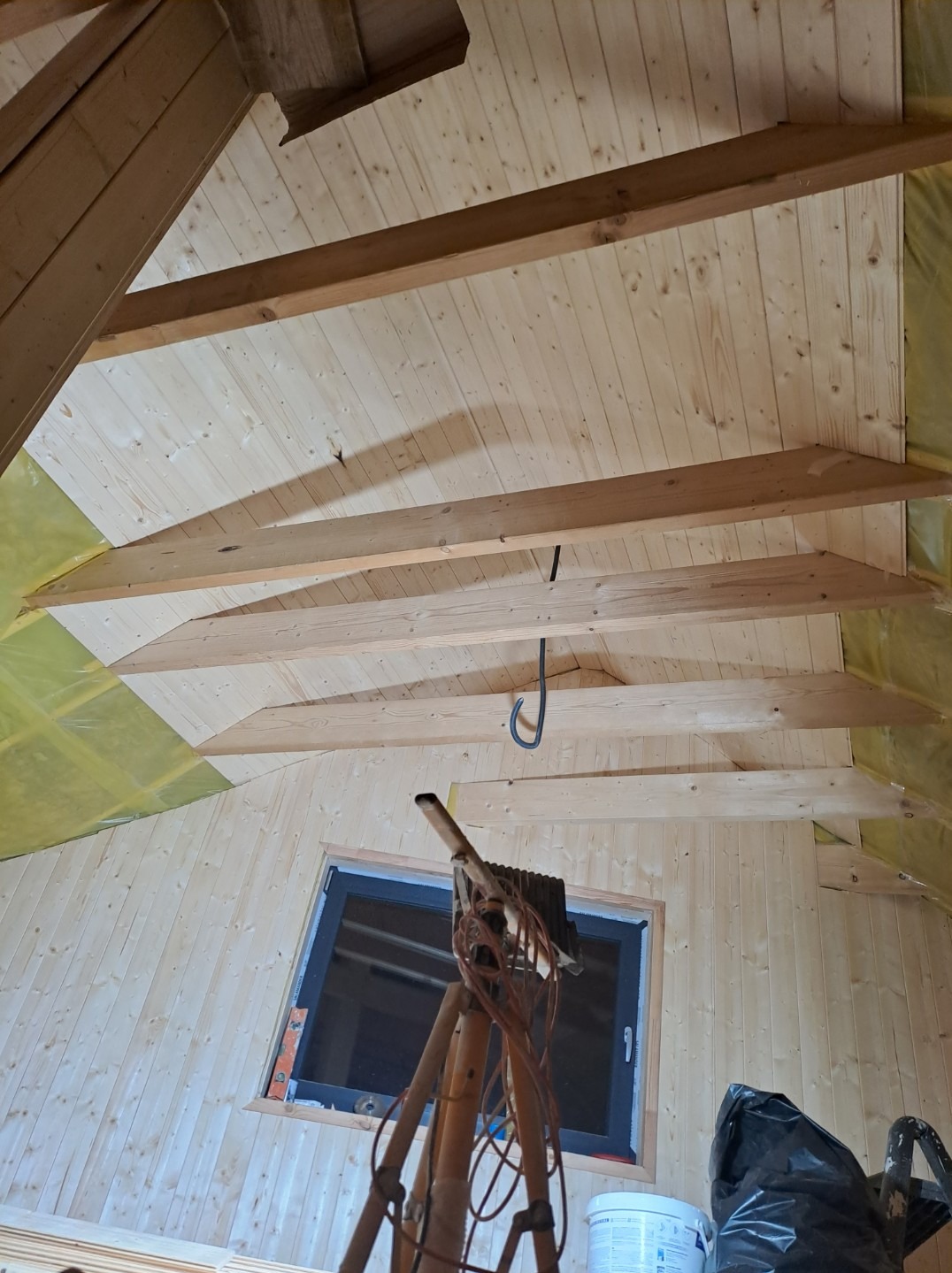 Wykończone drewnem poddasze ze skosem, widoczne drewniane belki stropowe, okno w szarej ramie, narzędzia i materiały budowlane na pierwszym planie, żółta folia izolacyjna w tle.