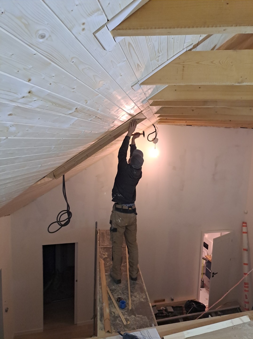 Montaż drewnianej boazerii na skośnym suficie poddasza przez pracownika stojącego na prowizorycznej drabinie, widoczne kable oświetleniowe i surowe ściany.