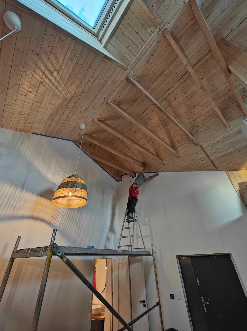 Pracownik na drabinie maluje ścianę przy drewnianym suficie z oknem dachowym, widoczne rusztowanie i wisząca lampa.