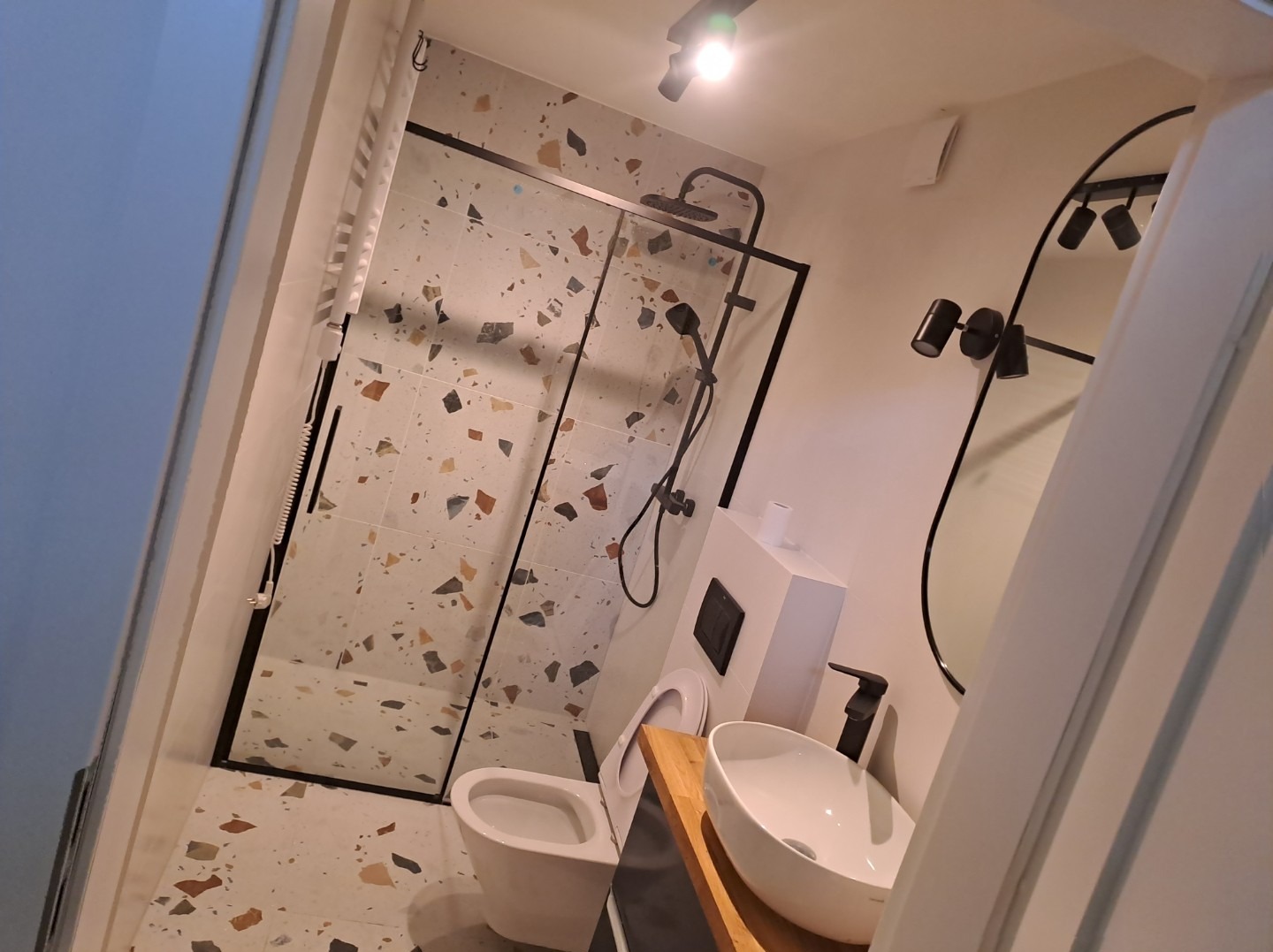 Nowoczesna łazienka z kabiną prysznicową typu walk-in z czarną ramą, umywalką nablatową na drewnianym blacie i lustrem z czarną ramą. Na ścianach i podłodze płytki w stylu lastryko.