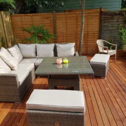 Słoneczne Ogrody - Umeblowany taras z drewna egzotycznego z narożną sofą rattanową, stolikiem kawowym ze szklanym blatem i pufami z jasnymi poduchami, otoczony drewnianym ogrodzeniem i zielenią.