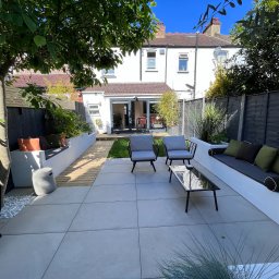 Słoneczne Ogrody - Nowoczesny taras z jasnych płyt, z białymi podwyższonymi rabatami i siedziskami z poduszkami, czarne krzesła i stolik kawowy, widok na zadbany trawnik i dom z przeszklonymi drzwiami tarasowymi.