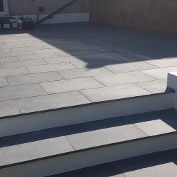 Słoneczne Ogrody - Nowoczesny taras z szarej kamiennej kostki, schody z kamienia prowadzące do domu, biała donica z ziemią w tle, ceglany mur.