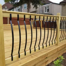 Nowy taras z jasnego drewna z czarnymi, metalowymi balustradami o falistym kształcie, widok z poziomu gruntu na tle domów jednorodzinnych.