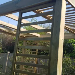 Surowa, drewniana pergola w trakcie montażu, widoczne elementy konstrukcyjne i jasne drewno na tle zieleni ogrodowej i zabudowań.