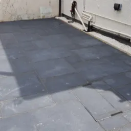 Nowoczesny taras wyłożony ciemnoszarymi płytami kamiennymi o zróżnicowanych wymiarach, z widoczną studzienką rewizyjną i obrzeżem z białego grysu przy ścianie budynku.