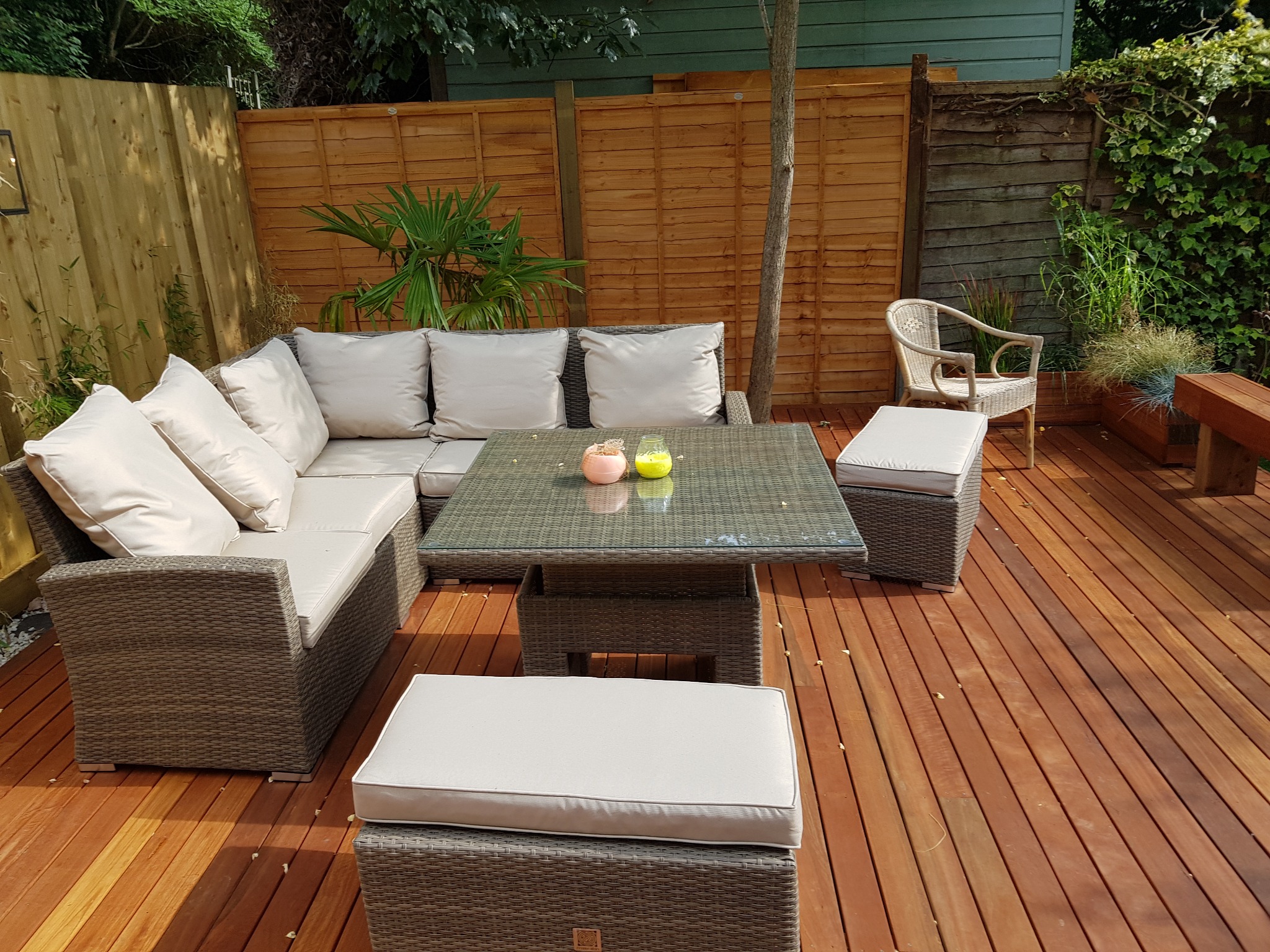 Umeblowany taras z drewna egzotycznego z narożną sofą rattanową, stolikiem kawowym ze szklanym blatem i pufami z jasnymi poduchami, otoczony drewnianym ogrodzeniem i zielenią.