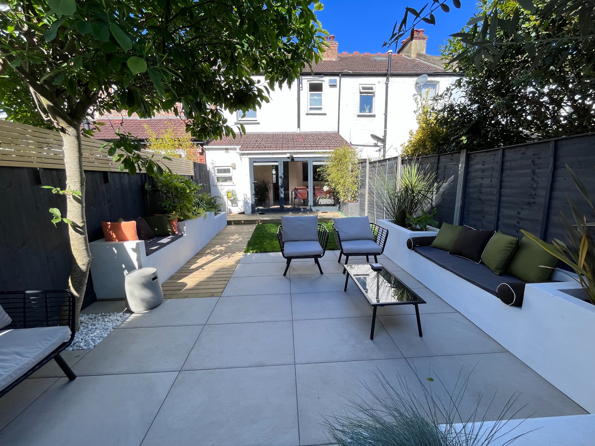 Nowoczesny taras z jasnych płyt, z białymi podwyższonymi rabatami i siedziskami z poduszkami, czarne krzesła i stolik kawowy, widok na zadbany trawnik i dom z przeszklonymi drzwiami tarasowymi.