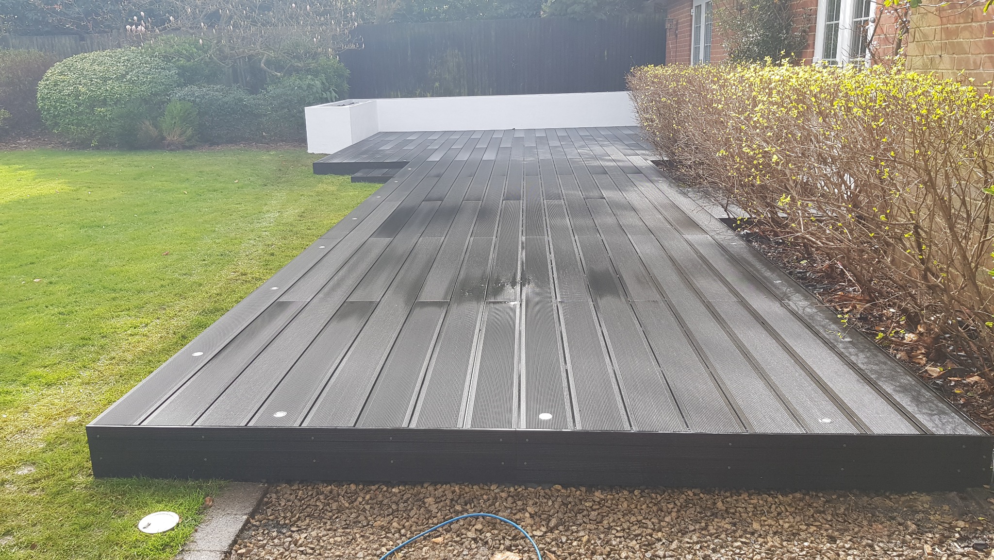 Nowoczesny taras z desek kompozytowych w kolorze grafitowym, z wbudowanym oświetleniem punktowym, przylegający do trawnika i krzewów w ogrodzie.