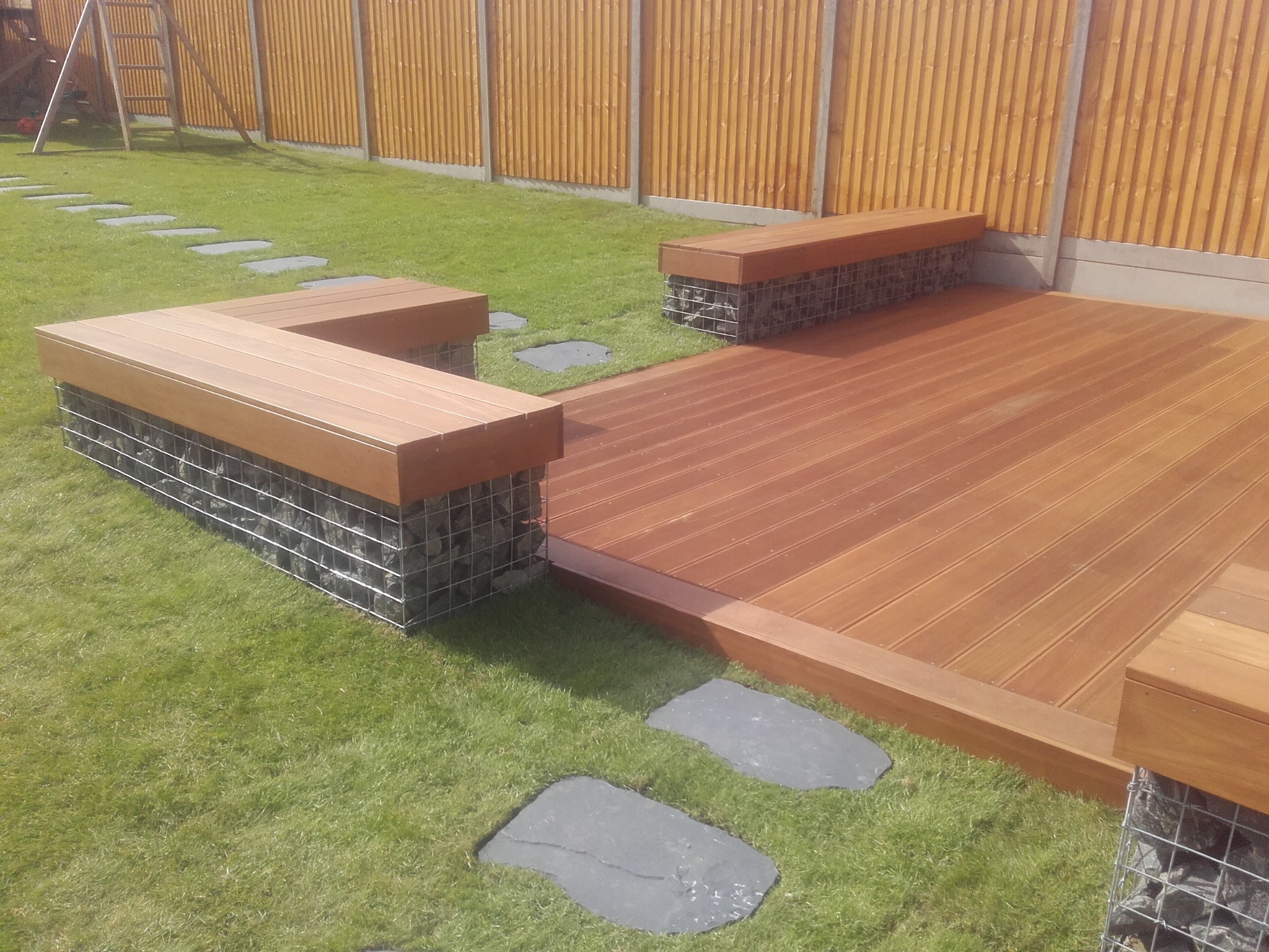 Nowoczesny taras z desek kompozytowych z siedziskami z gabionów i drewnianym blatem, otoczony trawnikiem z kamiennymi ścieżkami.