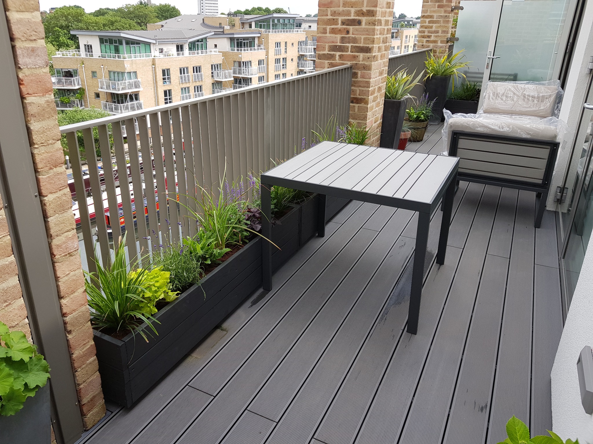 Nowoczesny taras na balkonie z szarą deską kompozytową, stolikiem, donicami z zielenią i meblami ogrodowymi pod folią ochronną, widok na budynki mieszkalne.